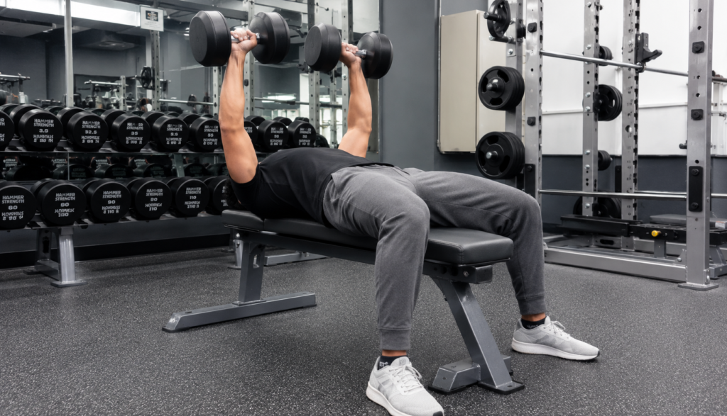 Person at lockout during dumbbell bench press in wide gym shot