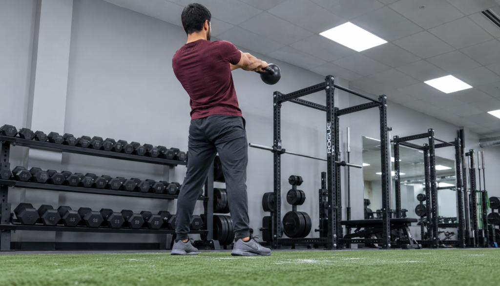 Lifter at the top of a kettlebell swing seen from behind on gym turf